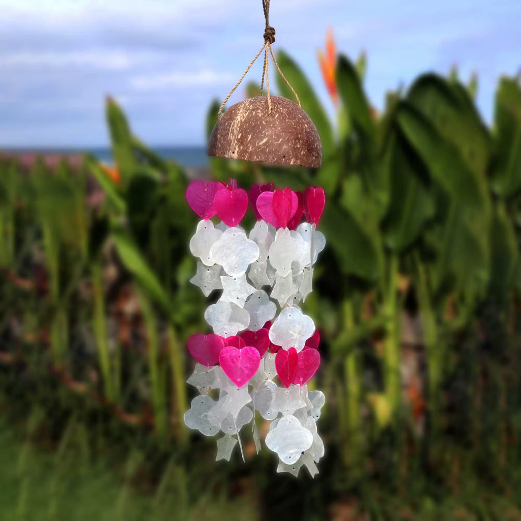 Coconut and capiz shell wind chime with pink heart and white flower shapes hanging in tropical garden
