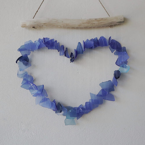 Blue glass heart wind chime on white wall — full heart shape made from mixed blue recycled glass pieces hanging from natural driftwood