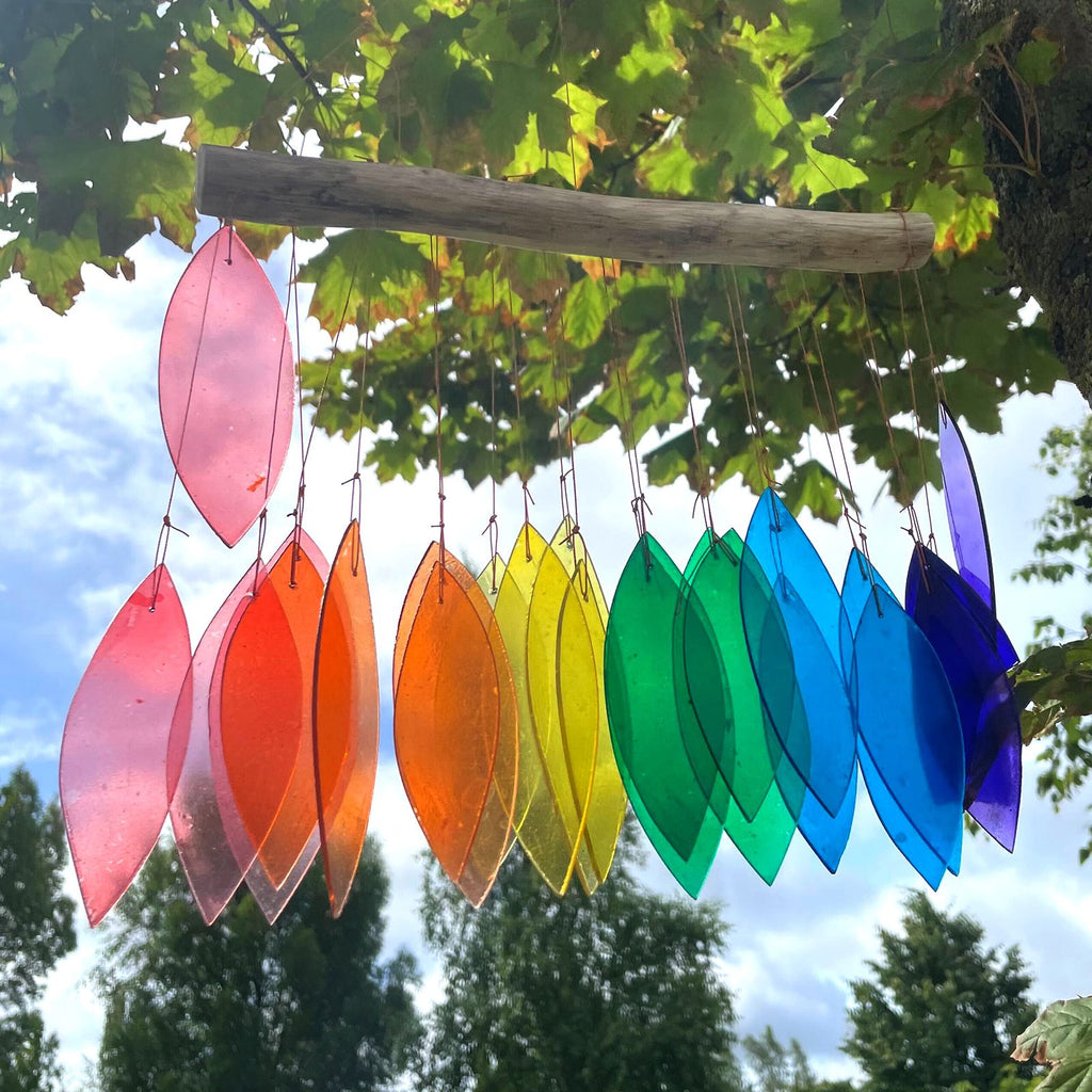 Rainbow glass wind chime hanging from tree branch in garden — leaf-shaped coloured glass pieces in pink, red, orange, yellow, green, teal, blue and purple