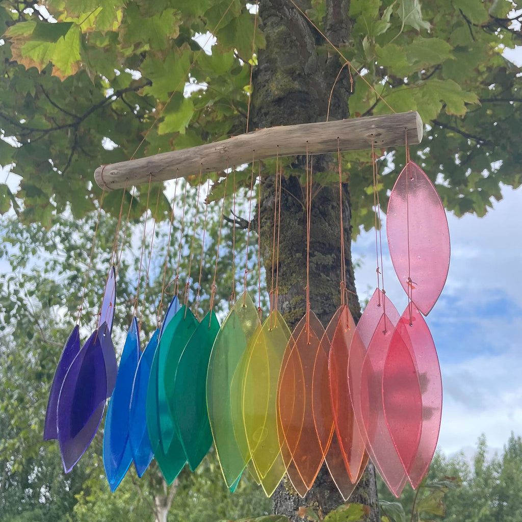 Rainbow glass wind chime hanging from tree — large leaf-shaped glass pieces in pink, red, orange, yellow, green, blue and purple catching light in garden setting