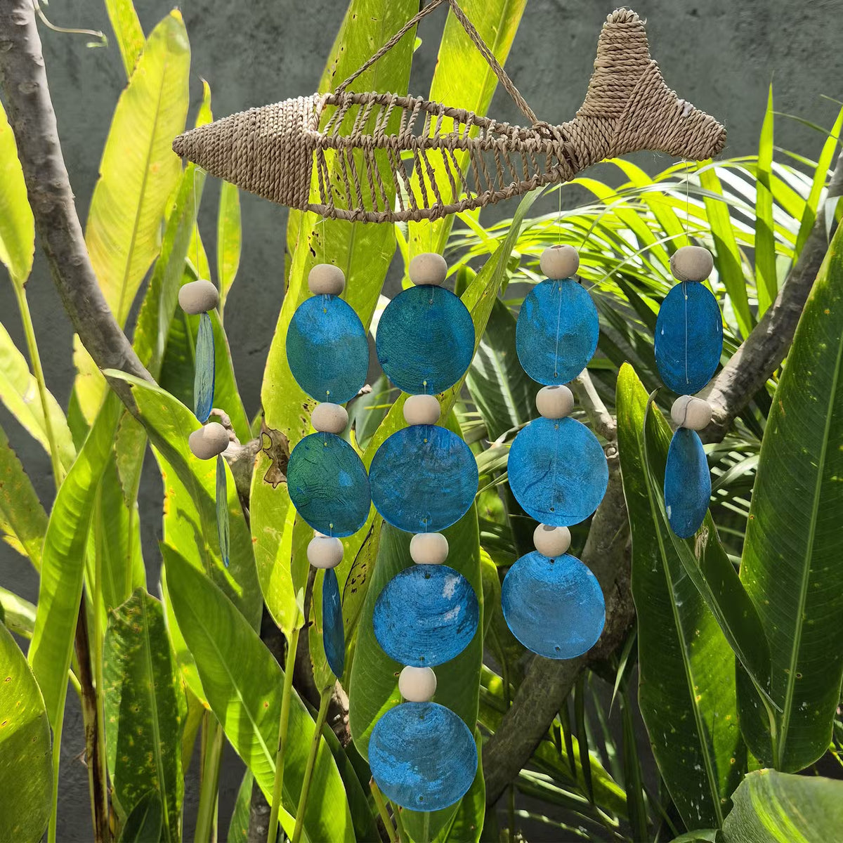 String fish woven decoration with blue capiz shell drops hanging among tropical foliage